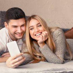 Loving young couple with mobile phone lying on sofa at home